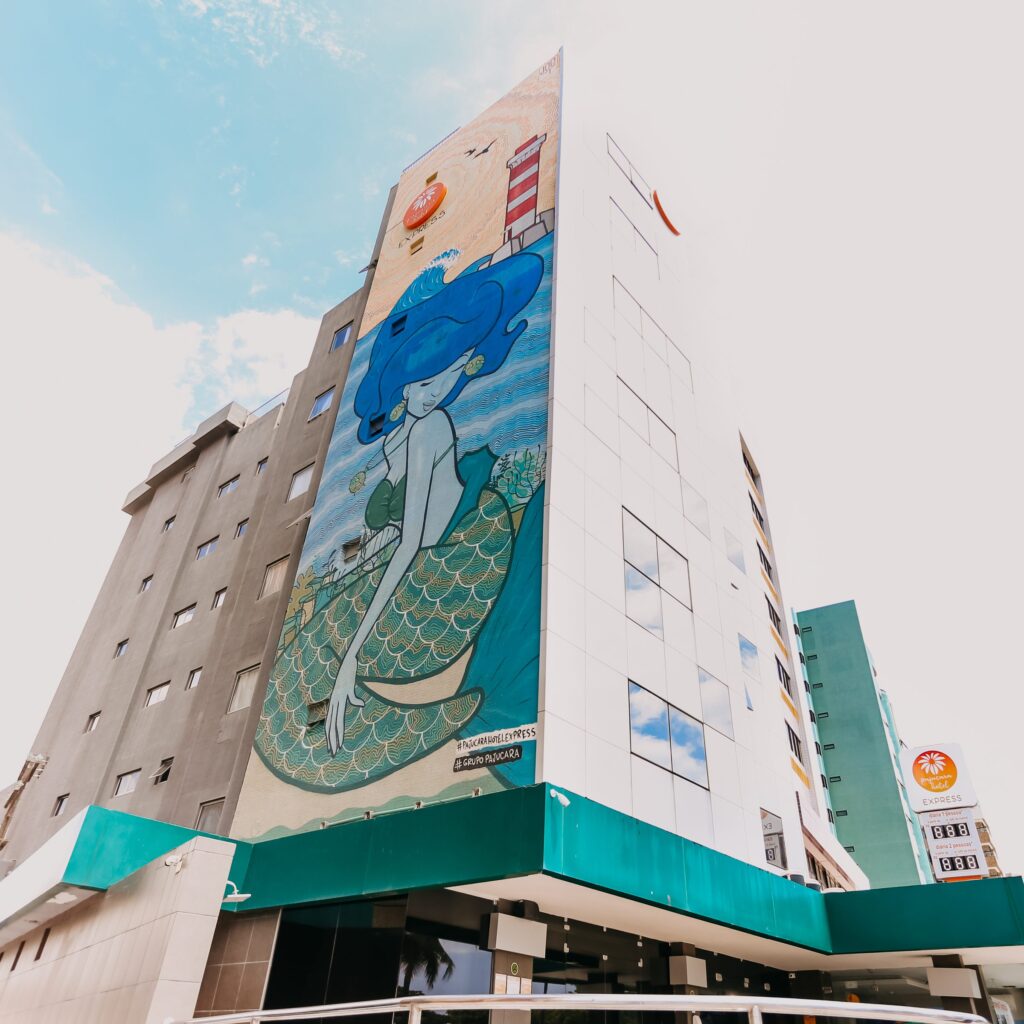 Fachada do hotel vista de baixo, com o painel artístico da sereia pintado na empena e o céu azul ao fundo