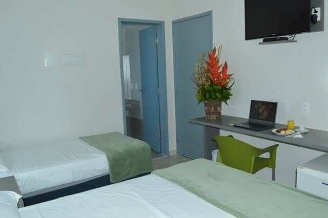 Quarto Lateral Mar com cama de casal e cama de solteiro, escrivaninha com cadeira, TV na parede e porta do banheiro