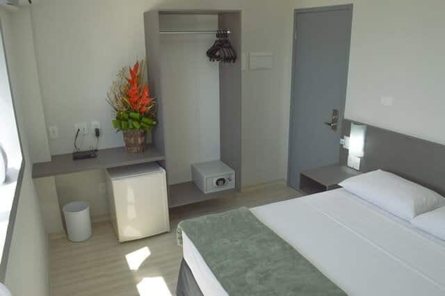 Quarto Lateral Mar com cama de casal, closet aberto com cabides, cofre, frigobar e arranjo de flores tropicais sobre a bancada
