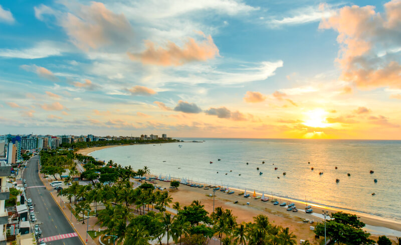 Vista aérea da orla da Pajuçara ao pôr do sol, com a praia, os coqueiros e o mar