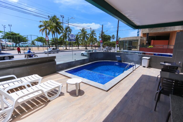 Piscina ao ar livre com espreguiçadeiras brancas e a orla da Pajuçara logo em frente