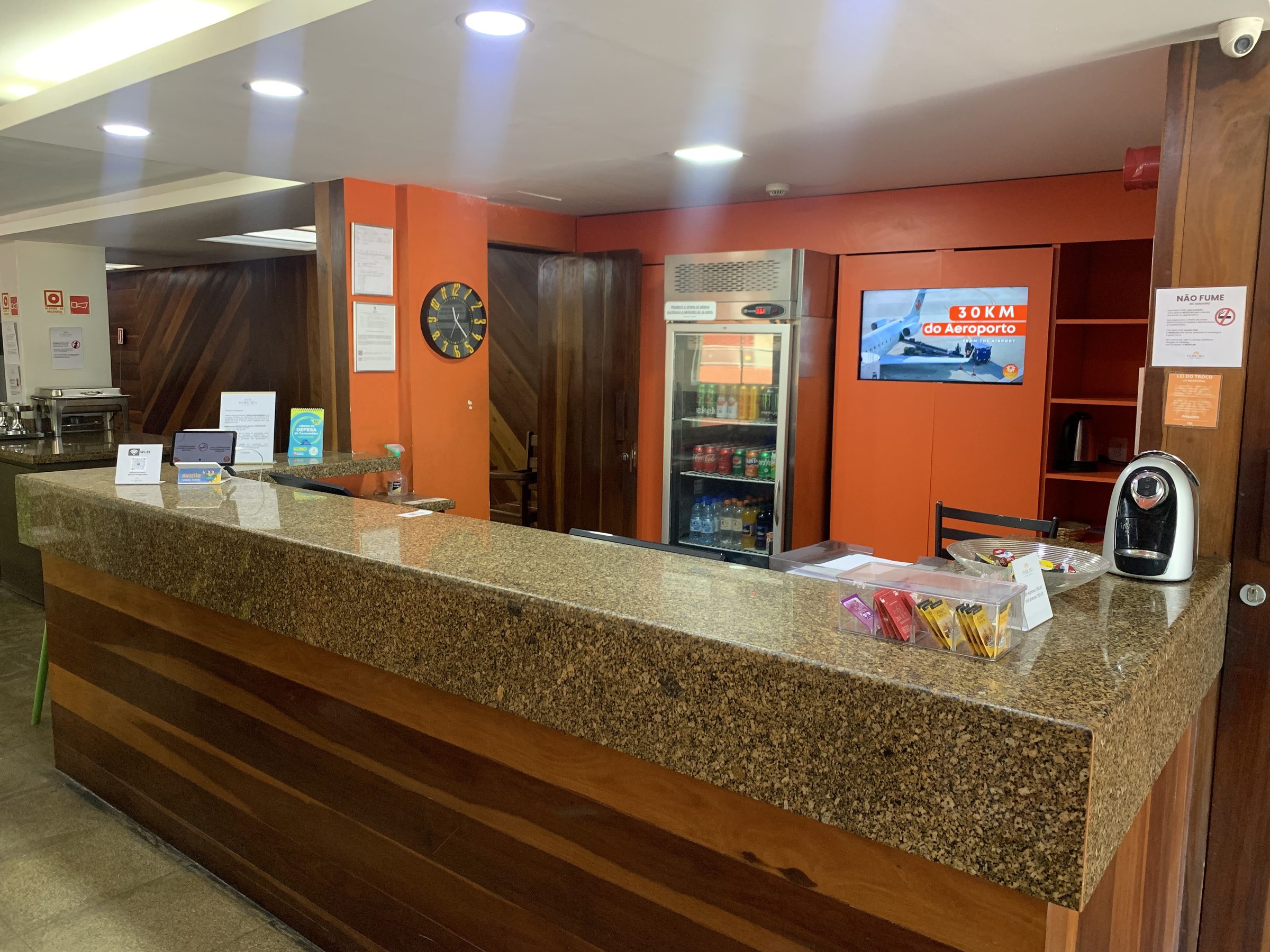 Balcão da recepção em madeira, com a lojinha de conveniência e o frigobar de bebidas ao fundo